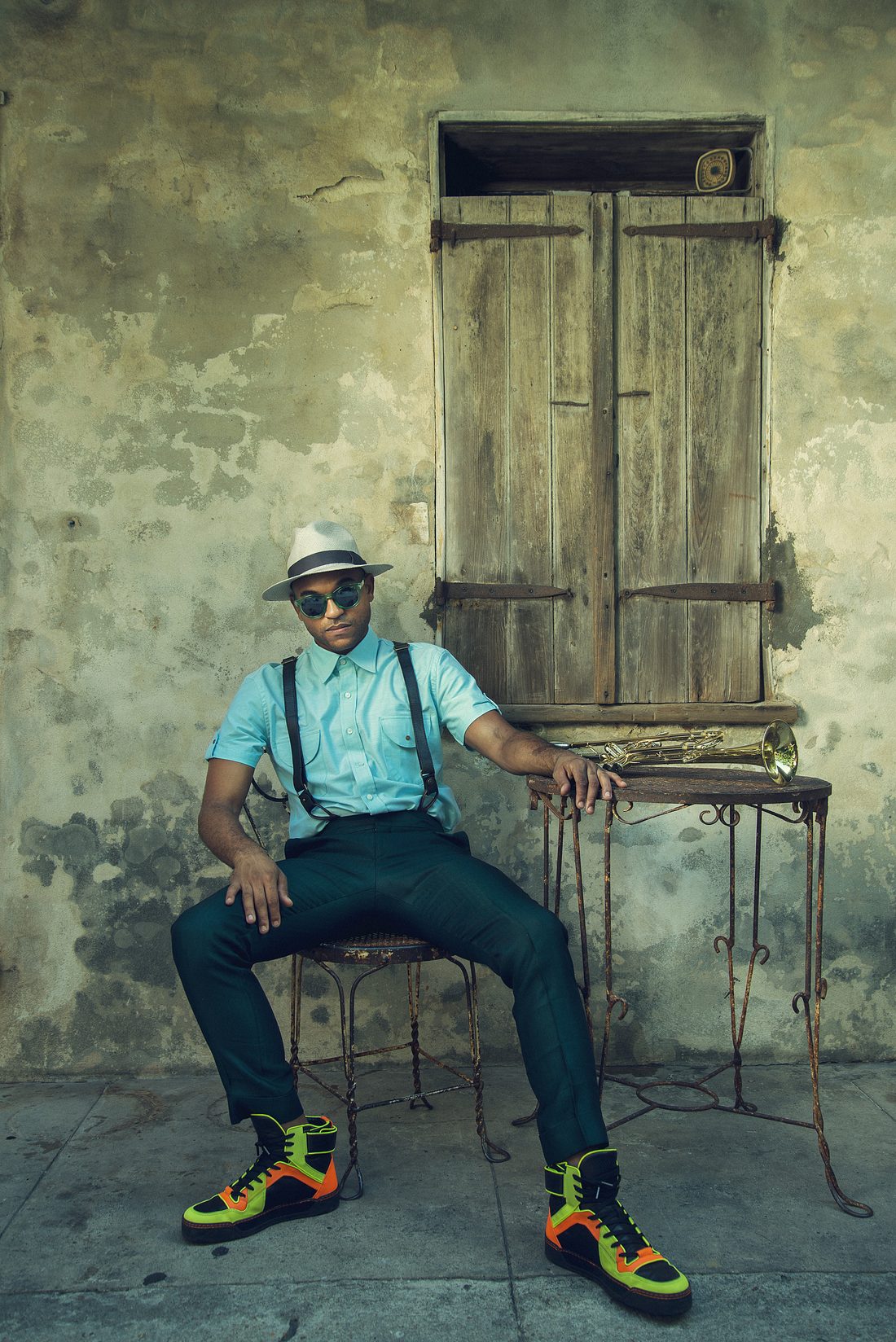 Branden Lewis in a straw hat and sunglasses, seated beside a trumpet on an iron table against a weathered stucco wall.