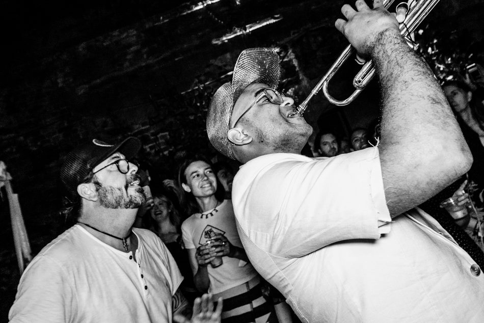 Black-and-white photograph of Branden Lewis in a flat cap, horn raised, playing in the middle of a packed crowd.