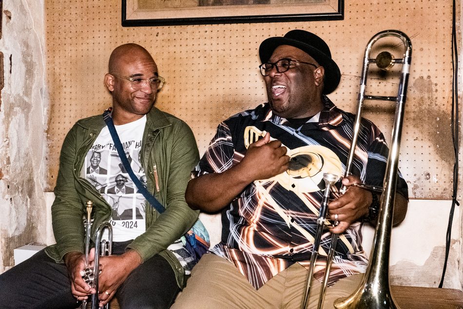 Branden Lewis, trumpet in hand, sitting on a bench beside Ronell Johnson, who holds his trombone mid-laugh, in a back room at Preservation Hall.