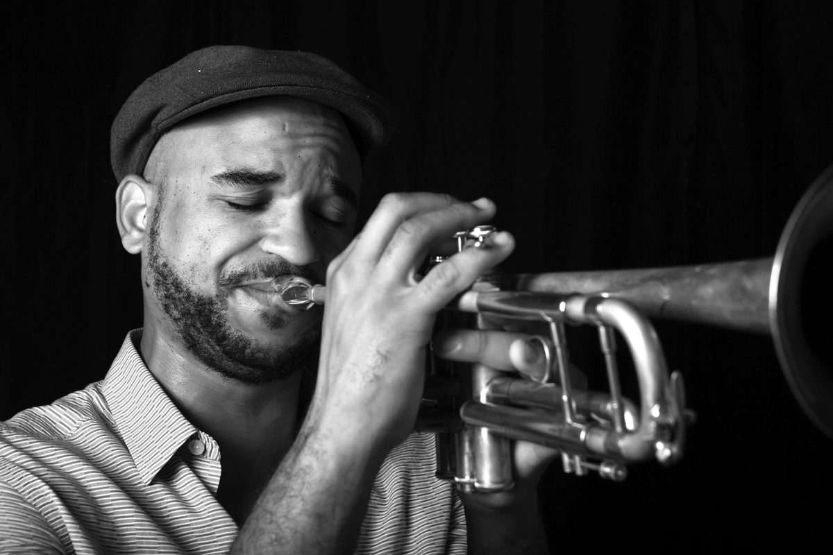 Black-and-white photograph of Branden Lewis mid-phrase on trumpet, wearing a flat cap.