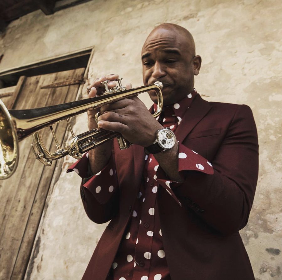 Branden Lewis playing trumpet in a burgundy suit and polka-dot shirt against a weathered wall.