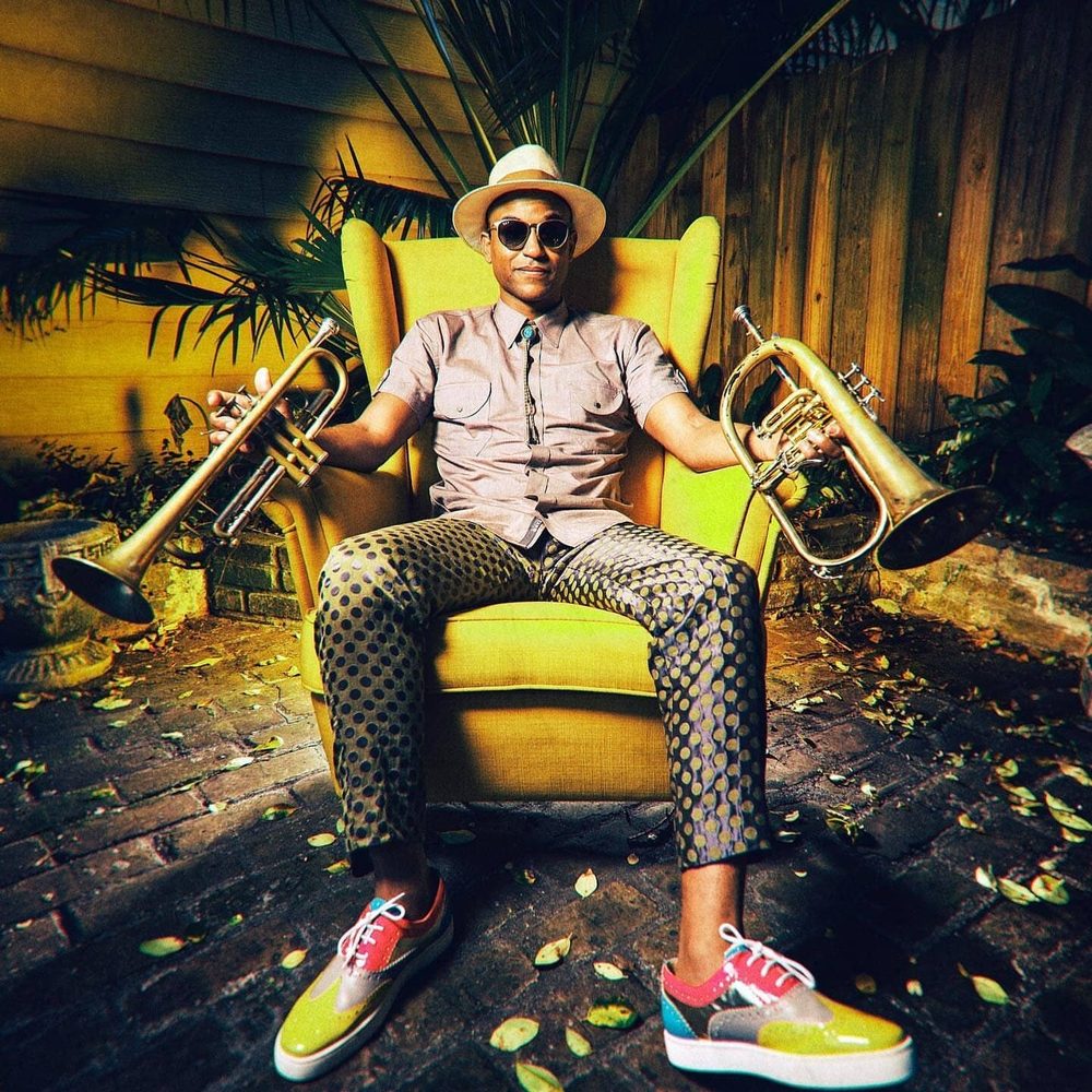 Branden Lewis seated in a yellow armchair in a courtyard, holding a trumpet in one hand and a flugelhorn in the other.